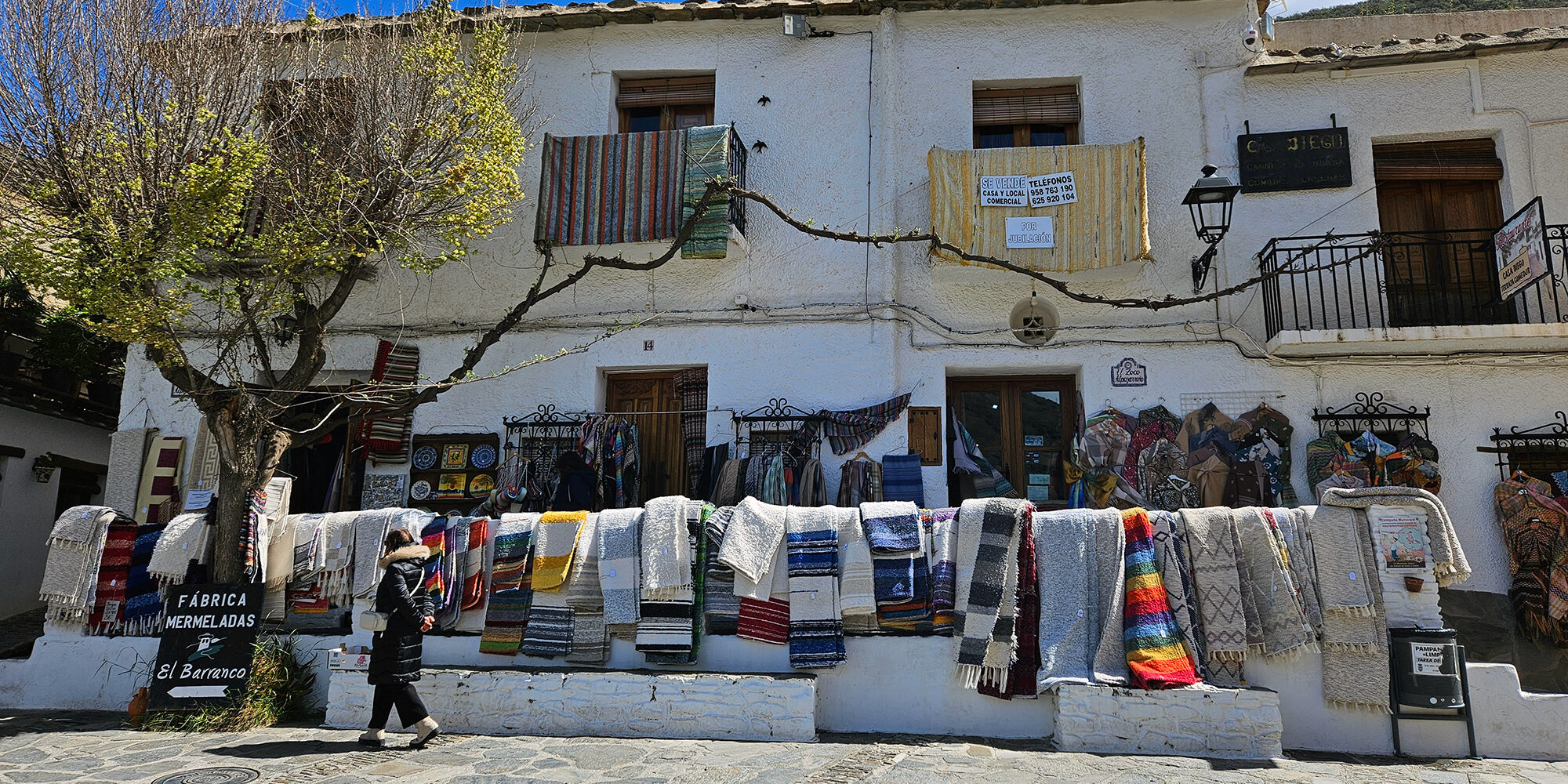 Pampaneira Plaza central de Pampaneira, Alpujarra de Granada: vista pintoresca de la plaza principal con una tienda tradicional de jarapas, donde se exhiben coloridos textiles artesanales. Un reflejo del encanto rústico y la autenticidad de este pintoresco pueblo blanco andaluz.