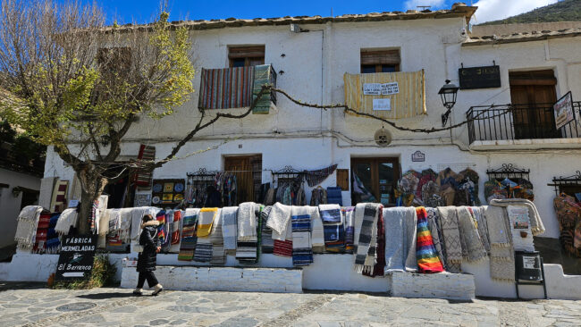 Plaza central de Pampaneira, Alpujarra de Granada: vista pintoresca de la plaza principal con una tienda tradicional de jarapas, donde se exhiben coloridos textiles artesanales. Un reflejo del encanto rústico y la autenticidad de este pintoresco pueblo blanco andaluz.