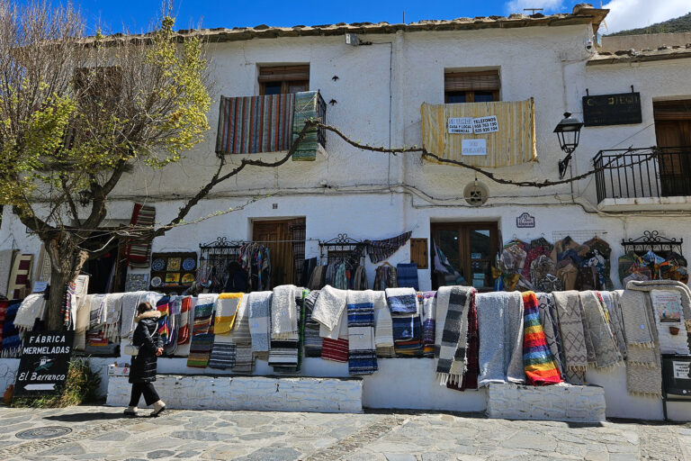 Plaza central de Pampaneira, Alpujarra de Granada: vista pintoresca de la plaza principal con una tienda tradicional de jarapas, donde se exhiben coloridos textiles artesanales. Un reflejo del encanto rústico y la autenticidad de este pintoresco pueblo blanco andaluz.